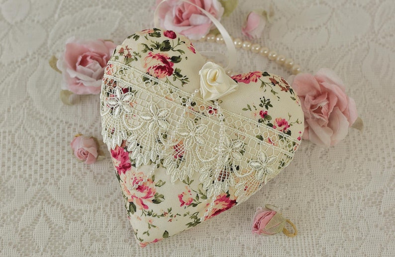 May include: A heart-shaped pillow with a floral print and lace trim. The pillow is on a white lace tablecloth with pink roses.