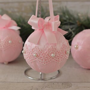 May include: Three pink Christmas baubles with white lace and pearl accents. The baubles are decorated with pink satin bows.