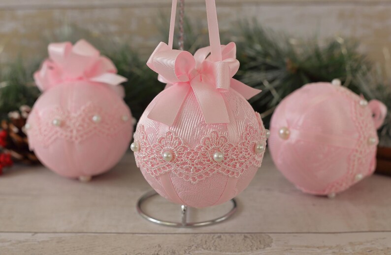 May include: Three pink Christmas baubles with white lace and pearl accents. The baubles are decorated with pink satin bows.