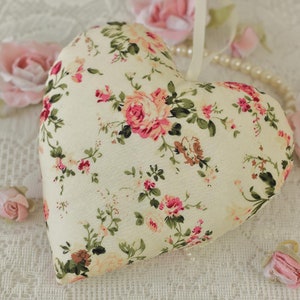 May include: A white heart-shaped pillow with a floral pattern of pink and green roses. The pillow is on a white lace background.