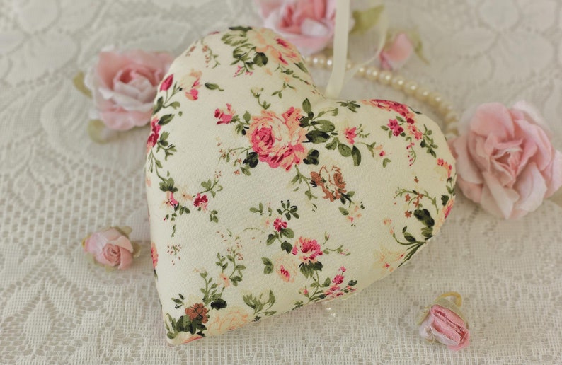 May include: A white heart-shaped pillow with a floral pattern of pink and green roses. The pillow is on a white lace background.