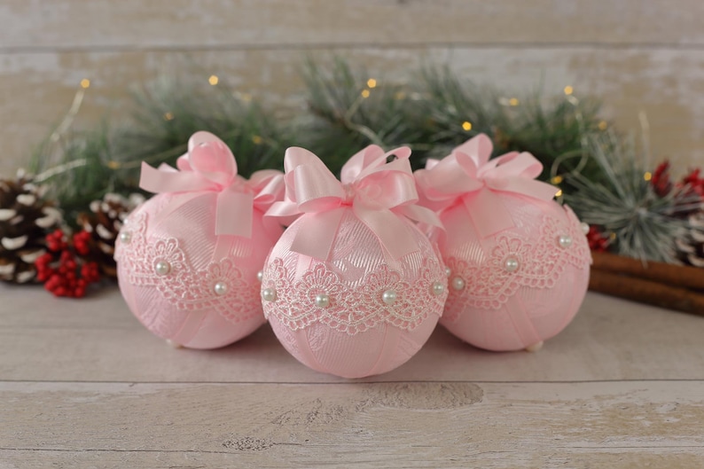 May include: Three pink Christmas baubles with white lace and pearl accents. Each bauble has a pink satin ribbon bow.