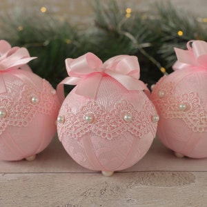 May include: Three pink Christmas baubles with white lace and pearl accents. Each bauble has a pink satin bow.