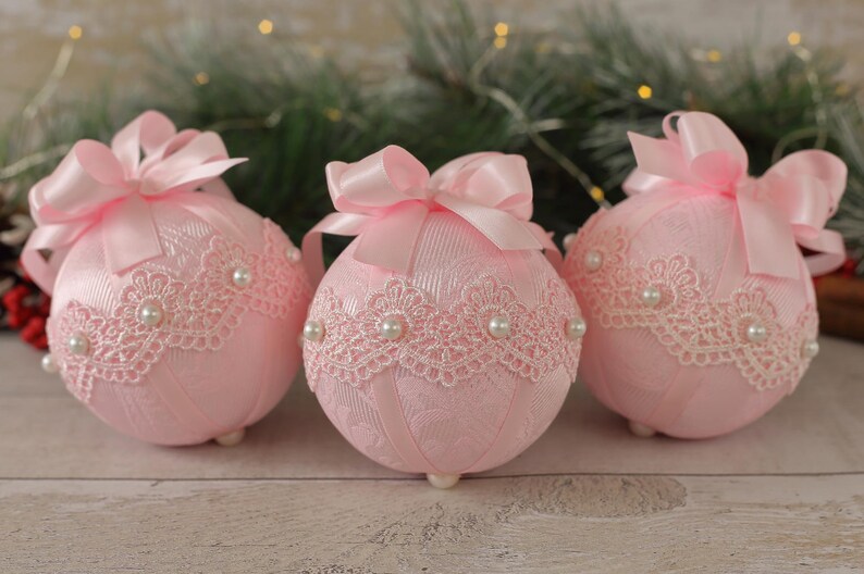 May include: Three pink Christmas baubles with white lace and pearl accents. Each bauble has a pink satin bow.