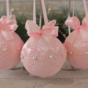 May include: Three pink Christmas baubles with white lace trim and pearl accents. Each bauble has a pink satin bow.