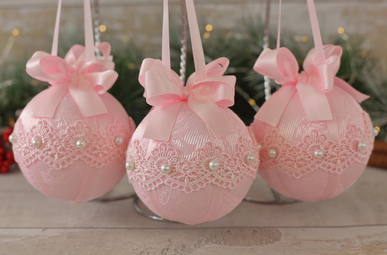 May include: Three pink Christmas baubles with white lace trim and pearl accents. Each bauble has a pink satin bow.