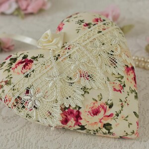 May include: A heart-shaped pillow with a floral print and lace trim. The pillow is cream-colored with pink and red roses. The pillow is sitting on a white lace tablecloth.