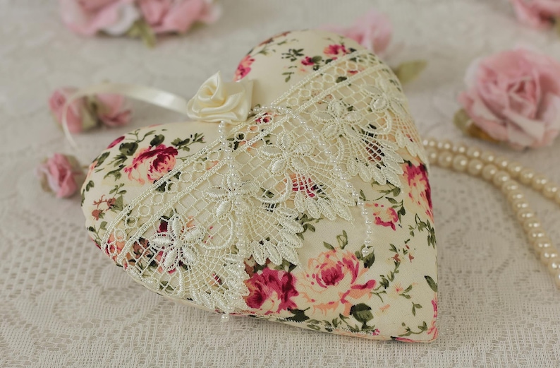 May include: A heart-shaped pillow with a floral print and lace trim. The pillow is cream-colored with pink and red roses. The pillow is sitting on a white lace tablecloth.