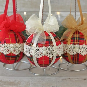 May include: Three red and white plaid Christmas ornaments with white lace trim and pearl accents. Each ornament is tied with a ribbon bow in a different color: red, white, and gold.
