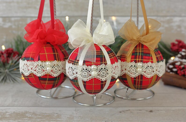 May include: Three red and white plaid Christmas ornaments with white lace trim and pearl accents. Each ornament is tied with a ribbon bow in a different color: red, white, and gold.