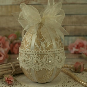 May include: A decorative Easter egg with a gold and white patterned design, adorned with a white lace trim and a white bow. The egg is sitting on a white lace doily.