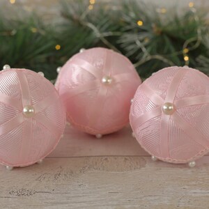 May include: Three pink Christmas baubles with white ribbon and pearl accents. The baubles are made of fabric and have a delicate, feminine design.