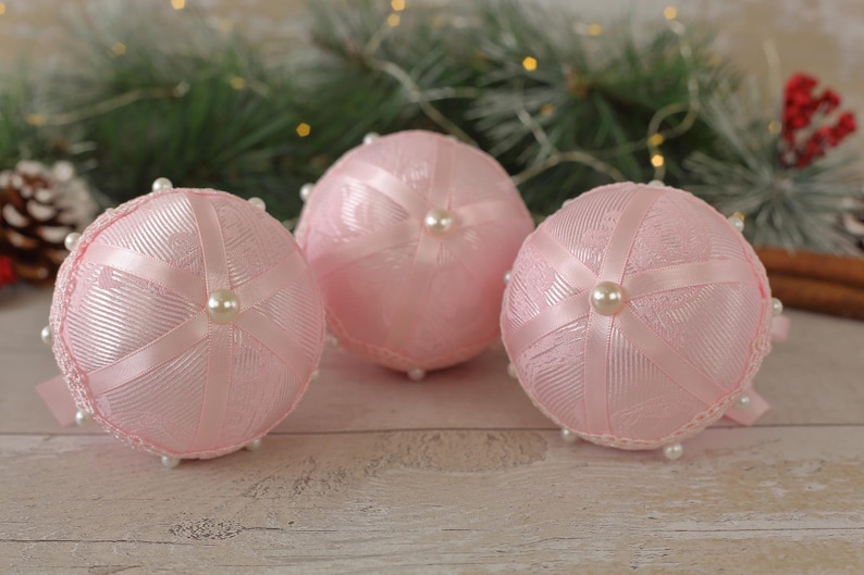 May include: Three pink Christmas baubles with white ribbon and pearl accents. The baubles are made of fabric and have a delicate, feminine design.