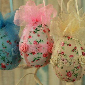 Peut inclure: Trois ornements d&eacute;coratifs en forme d'&oelig;ufs de P&acirc;ques. Chaque &oelig;uf est recouvert d'un tissu floral avec une garniture en ruban et en dentelle. Les &oelig;ufs sont orn&eacute;s de n&oelig;uds bleus, roses et jaunes, et de petites roses en tissu.