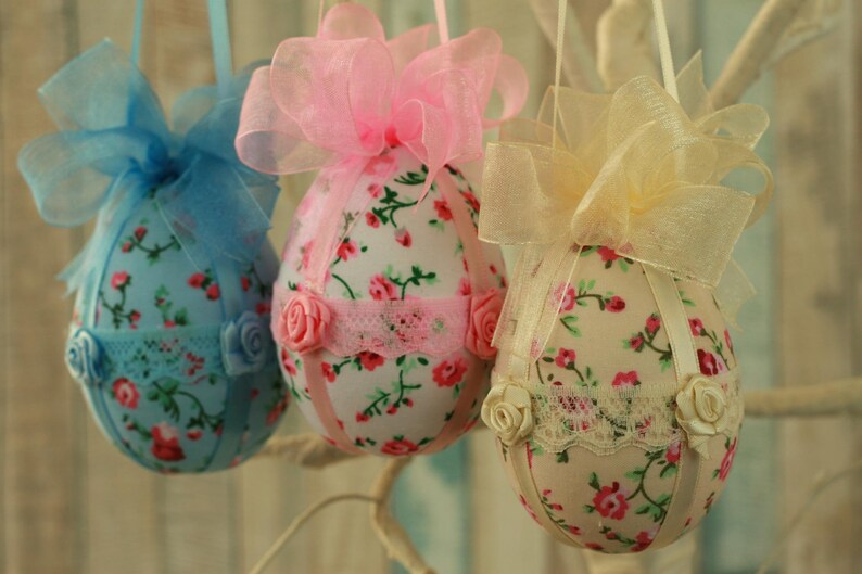 Peut inclure: Trois ornements d&eacute;coratifs en forme d'&oelig;ufs de P&acirc;ques. Chaque &oelig;uf est recouvert d'un tissu floral avec une garniture en ruban et en dentelle. Les &oelig;ufs sont orn&eacute;s de n&oelig;uds bleus, roses et jaunes, et de petites roses en tissu.