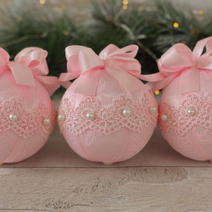 May include: Three pink Christmas baubles with white lace trim and pearl accents. Each bauble has a pink satin bow.