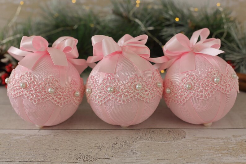May include: Three pink Christmas baubles with white lace trim and pearl accents. Each bauble has a pink satin bow.