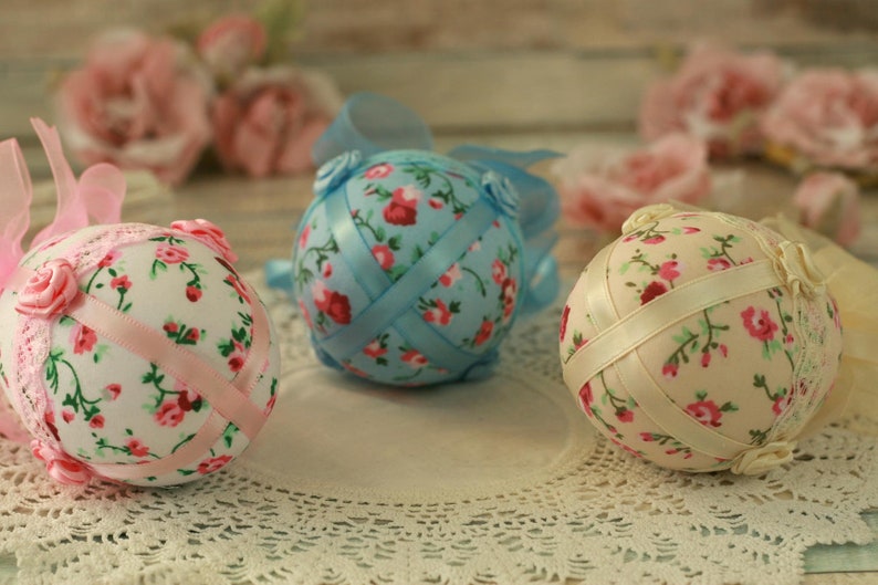 Peut inclure: Trois sph&egrave;res d&eacute;coratives florales avec des rubans. Une sph&egrave;re est rose clair avec un ruban rose, une autre est bleu clair avec un ruban bleu, et la troisi&egrave;me est cr&egrave;me avec un ruban cr&egrave;me. Chaque sph&egrave;re a un motif floral avec des roses rouges et roses.