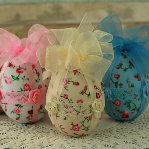 Peut inclure: Trois &oelig;ufs de P&acirc;ques d&eacute;coratifs, chacun orn&eacute; d'un tissu floral et d'un n&oelig;ud en ruban transparent. Les &oelig;ufs pr&eacute;sentent des accents roses, jaunes et bleus, avec de la dentelle et des roses. Parfaits pour les d&eacute;corations de P&acirc;ques.