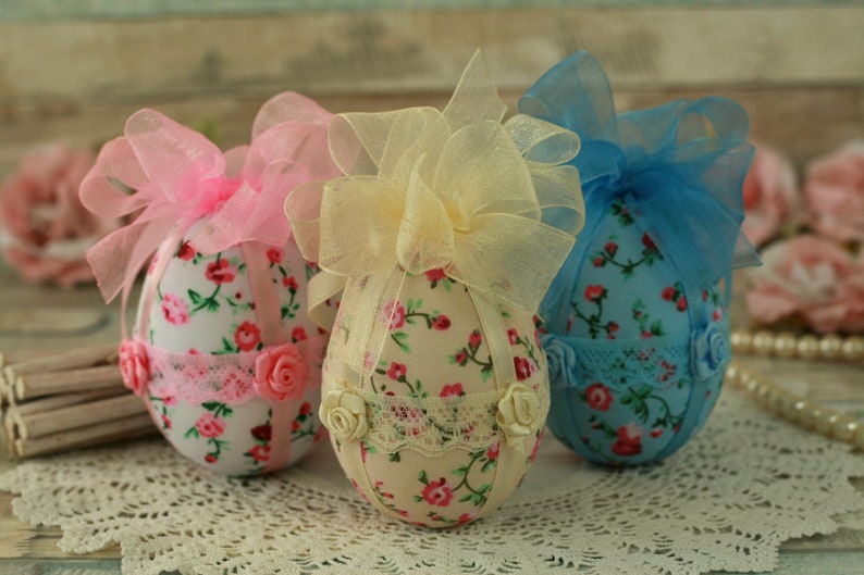 Peut inclure: Trois &oelig;ufs de P&acirc;ques d&eacute;coratifs, chacun orn&eacute; d'un tissu floral et d'un n&oelig;ud en ruban transparent. Les &oelig;ufs pr&eacute;sentent des accents roses, jaunes et bleus, avec de la dentelle et des roses. Parfaits pour les d&eacute;corations de P&acirc;ques.