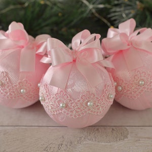 May include: Three pink Christmas baubles with white lace and pearl embellishments. Each bauble has a pink satin ribbon tied into a bow.