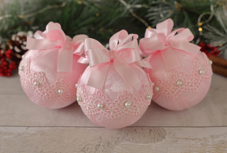 May include: Three pink Christmas baubles with white lace and pearl embellishments. Each bauble has a pink satin ribbon tied into a bow.