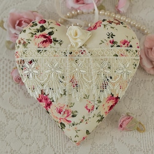 May include: A heart-shaped pillow with a floral print and lace trim. The pillow is decorated with a white satin rose and is sitting on a white lace tablecloth.