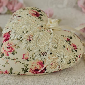 May include: A heart-shaped pillow with a floral print and lace trim. The pillow is decorated with a satin ribbon and pearls. The pillow is sitting on a white lace tablecloth.