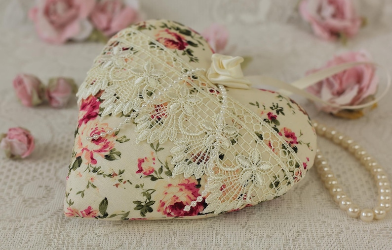 May include: A heart-shaped pillow with a floral print and lace trim. The pillow is decorated with a satin ribbon and pearls. The pillow is sitting on a white lace tablecloth.