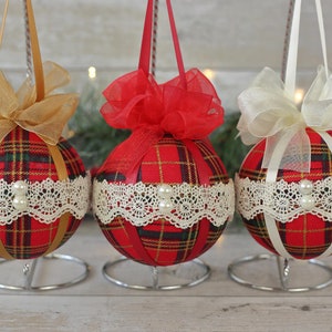 May include: Three red and black plaid Christmas ornaments with white lace trim and pearl accents. Each ornament has a different colored ribbon bow: gold, red, and white.