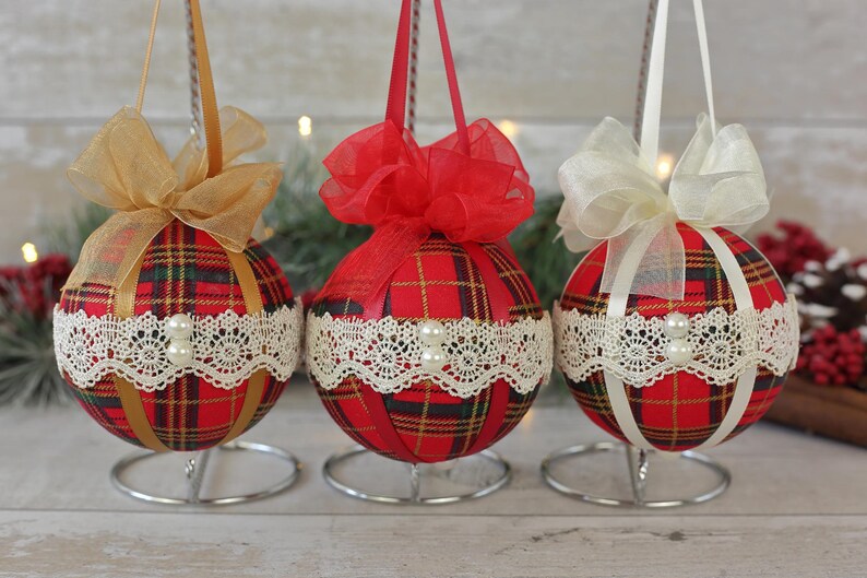 May include: Three red and black plaid Christmas ornaments with white lace trim and pearl accents. Each ornament has a different colored ribbon bow: gold, red, and white.