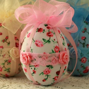 Peut inclure: Trois &oelig;ufs de P&acirc;ques d&eacute;coratifs en tissu. Les &oelig;ufs sont recouverts de motifs floraux avec des accents roses, rouges et verts. Chaque &oelig;uf a un n&oelig;ud en ruban en haut et est orn&eacute; de dentelle et de roses.