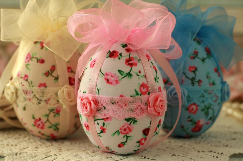 Peut inclure: Trois &oelig;ufs de P&acirc;ques d&eacute;coratifs en tissu. Les &oelig;ufs sont recouverts de motifs floraux avec des accents roses, rouges et verts. Chaque &oelig;uf a un n&oelig;ud en ruban en haut et est orn&eacute; de dentelle et de roses.