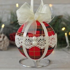 May include: A red and white plaid Christmas ornament with a white lace trim and a white satin ribbon bow. The ornament is on a silver stand.