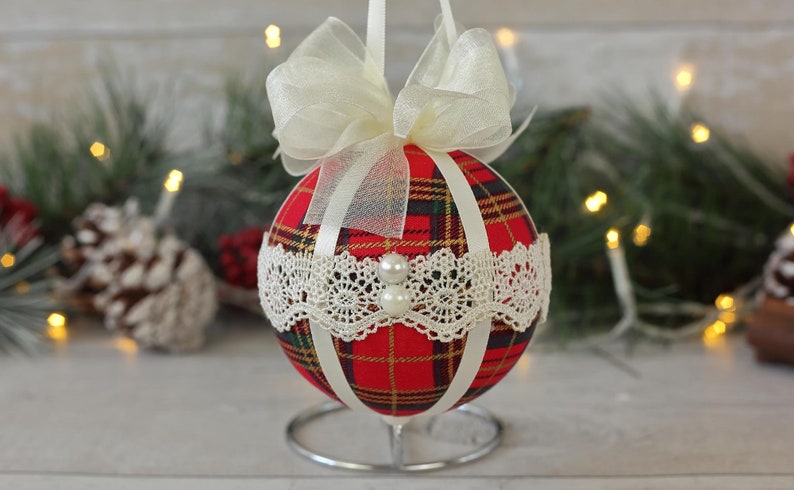 May include: A red and white plaid Christmas ornament with a white lace trim and a white satin ribbon bow. The ornament is on a silver stand.