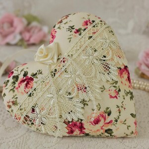 May include: A heart-shaped pillow with a floral print and lace trim. The pillow is cream-colored with pink and red roses. It is sitting on a white lace tablecloth.