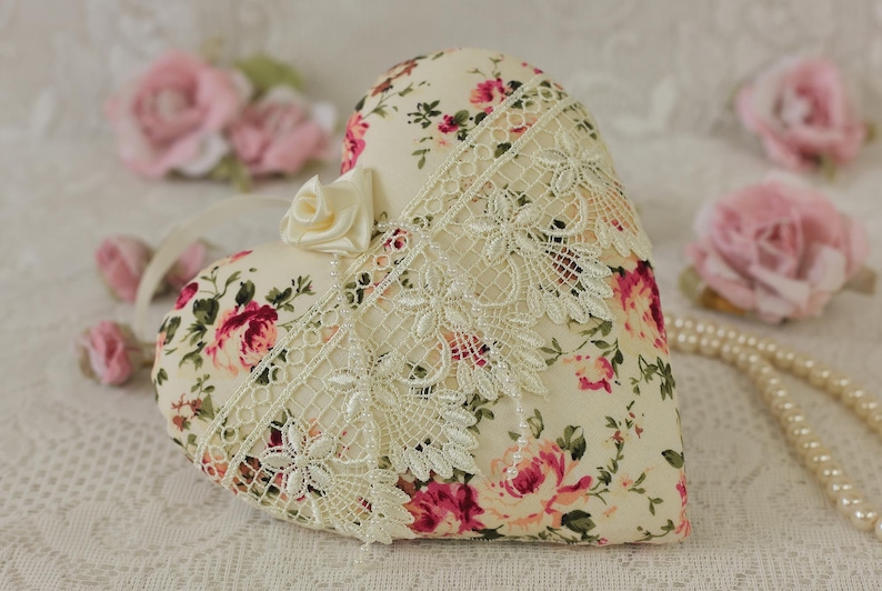 May include: A heart-shaped pillow with a floral print and lace trim. The pillow is cream-colored with pink and red roses. It is sitting on a white lace tablecloth.