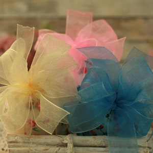 Peut inclure: Trois n&oelig;uds d&eacute;coratifs aux couleurs pastel : jaune, rose et bleu. Les n&oelig;uds sont faits de tissu transparent et d&eacute;licat, dispos&eacute;s dans un contenant en bois blanc rustique. L'arri&egrave;re-plan est flou.