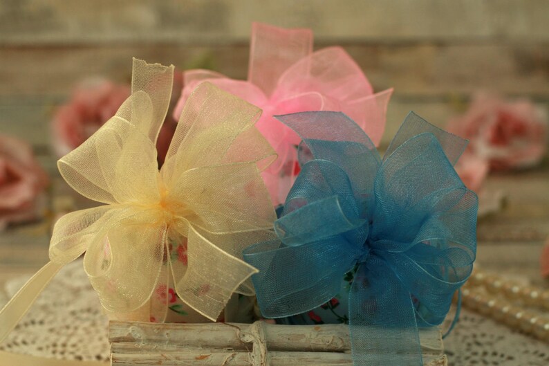 Peut inclure: Trois n&oelig;uds d&eacute;coratifs aux couleurs pastel : jaune, rose et bleu. Les n&oelig;uds sont faits de tissu transparent et d&eacute;licat, dispos&eacute;s dans un contenant en bois blanc rustique. L'arri&egrave;re-plan est flou.