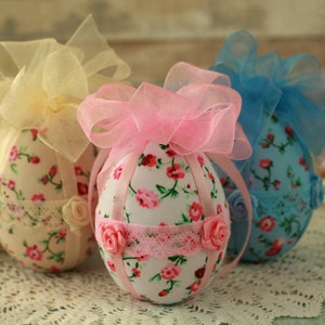 Peut inclure: Trois ornements d&eacute;coratifs en forme d'&oelig;uf. Chaque &oelig;uf est recouvert d'un tissu floral avec des fleurs roses et rouges sur fond blanc. Ils sont orn&eacute;s de dentelle, de ruban et de petites roses. Les n&oelig;uds sur le dessus sont jaunes, roses et bleus.