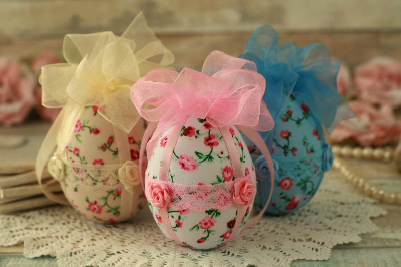 Peut inclure: Trois ornements d&eacute;coratifs en forme d'&oelig;uf. Chaque &oelig;uf est recouvert d'un tissu floral avec des fleurs roses et rouges sur fond blanc. Ils sont orn&eacute;s de dentelle, de ruban et de petites roses. Les n&oelig;uds sur le dessus sont jaunes, roses et bleus.