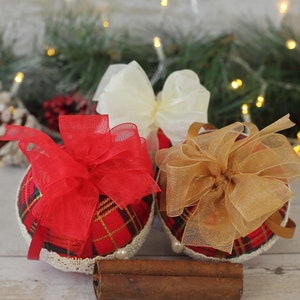 May include: Three Christmas ornaments with red, gold, and white bows. The ornaments are covered in a plaid fabric and have a white lace trim. The ornaments are sitting on a wooden surface with cinnamon sticks.
