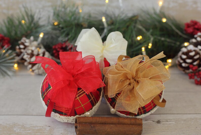 May include: Three Christmas ornaments with red, gold, and white bows. The ornaments are covered in a plaid fabric and have a white lace trim. The ornaments are sitting on a wooden surface with cinnamon sticks.
