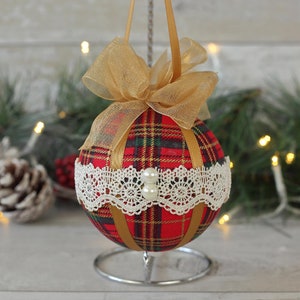 May include: A red and green plaid Christmas ornament with gold ribbon and white lace trim. The ornament is on a silver stand.
