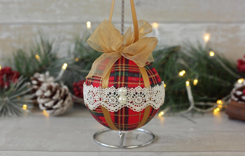 May include: A red and green plaid Christmas ornament with gold ribbon and white lace trim. The ornament is on a silver stand.