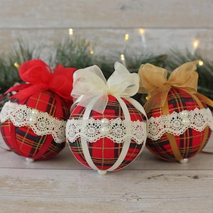 May include: Three red, white, and green plaid Christmas ornaments with white lace trim and pearl accents. Each ornament has a different colored ribbon bow.