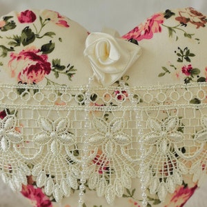 May include: A heart-shaped pillow with a floral print and a white lace trim. The pillow is decorated with a white satin rose.