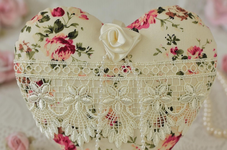 May include: A heart-shaped pillow with a floral print and a white lace trim. The pillow is decorated with a white satin rose.