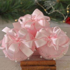 May include: Three pink Christmas baubles with lace and satin ribbon bows. The baubles are arranged on a wooden surface with cinnamon sticks.