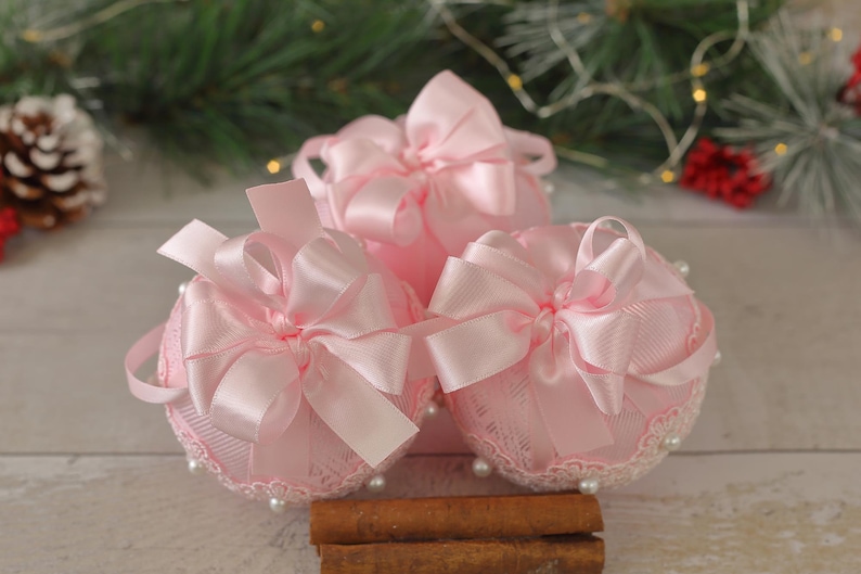 May include: Three pink Christmas baubles with lace and satin ribbon bows. The baubles are arranged on a wooden surface with cinnamon sticks.